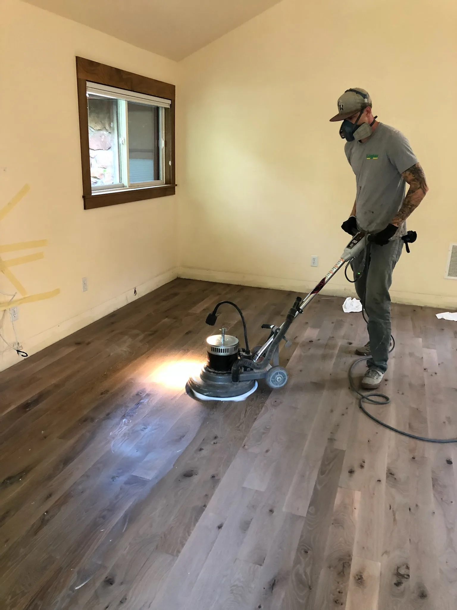 Flooring Craftsman Buffing A Dark-Stained Hardwood Floor With A Rotary Sander During Refinishing, Wearing A Respirator Mask In A Sparsely Furnished Room.