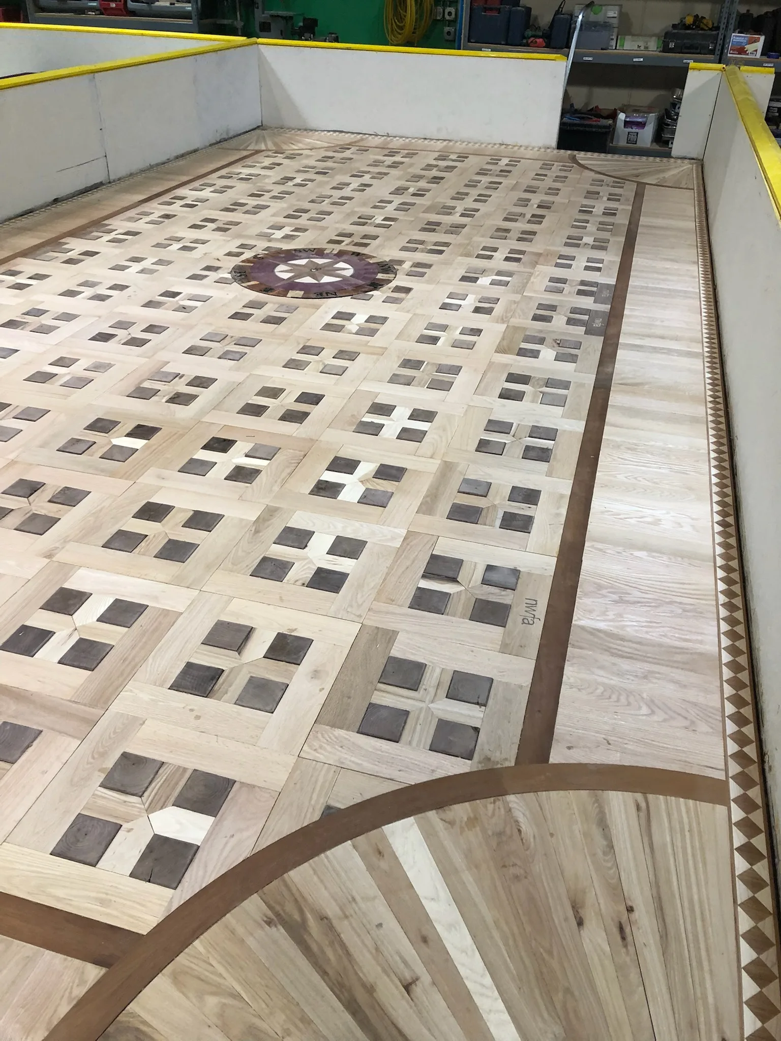 Custom Parquet Hardwood Floor With Light And Dark Wood Squares, Center Medallion, And Detailed Border Inlays Installed Inside An Indoor Rink-Style Room.